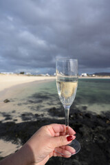 Hand with glass of white champagne or cava sparkling wine on white sandy tropical beach and blue ocean, romantic vacation, winter sun on Fuerteventura, Canary, Spain