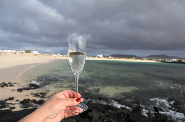 Hand with glass of white champagne or cava sparkling wine on white sandy tropical beach and blue ocean, romantic vacation, winter sun on Fuerteventura, Canary, Spain