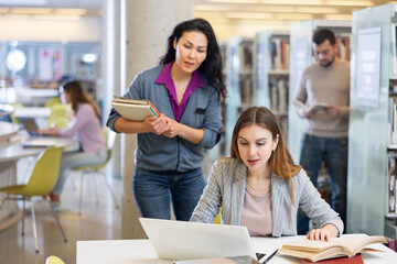 Female student working on laptop computer in library, reading books and making notes, preparing to exam
