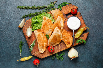 Plakat Wooden board with tasty meat empanadas, vegetables, herbs and sauce on dark background