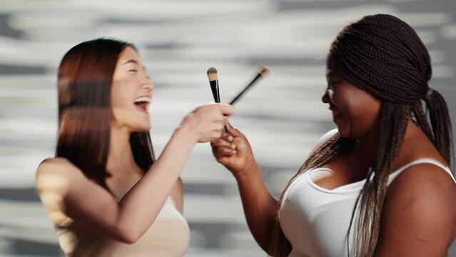 Happy multiracial girls playing with brushes in studio, giving eachother makeovers with cosmetics and having fun. Young beautiful models enjoying femininity and body positivity campaign.