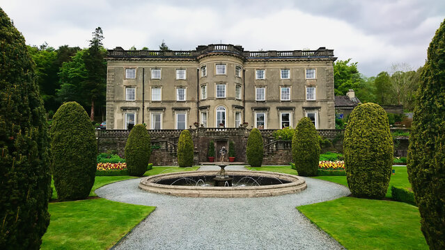 Rydal Mount and Gardens, Lake District, United Kingdom - May 11, 2018: front view of the exterior of the facade of the garden and house with a fountain in the front under a cloudy sky