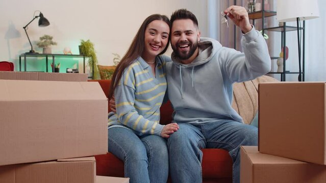 Young family marriage couple man woman lifting hand showing the keys of new home house apartment, buying or renting real estate property, mortgage loan at home. Husband wife together on sofa in room