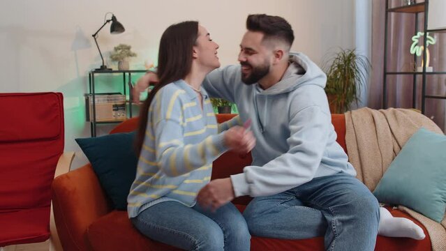 Young family married couple man woman at home on sofa in living room. Wife shows pregnancy positive test result to happy smiling husband. Celebrate, hug embrace. Excited newlyweds waiting future child - Powered by Adobe