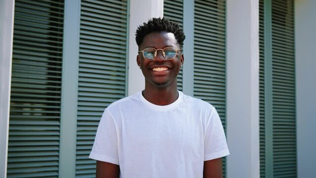 Portrait of one young african american teenager smiling and looking at camera standing outdoors. Front view of happy and joyful man with eyeglasses and white perfect teeth. slow motion. High quality