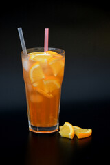 Tall faceted glass of refreshing lemonade with ice, straws and orange slices on a black background.