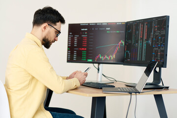 Serious handsome trader holding mobile phone, using laptop computer working online with crypto chart sitting in modern office. Successful business, online trading concept  