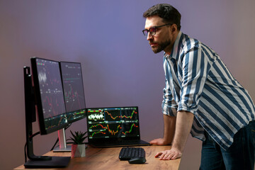 Night portrait of serious crypto trader looking at computer monitor, working online with crypto chart standing in modern office, researching and analyzing market. Technology, online trading concept