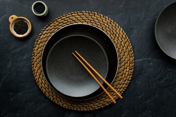 A table setting on a straw mat, with chopsticks, soya sauce and sesame seeds.