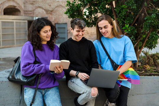 Young diverse people with lgbt rainbow flag using laptop and book outdoors.