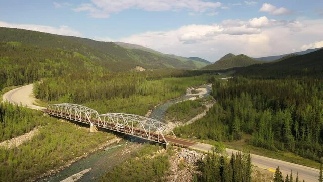 Steel bridge on the Canadian section of the Alaska Highway. Summer season, sunny day. Road, forested mountains, river