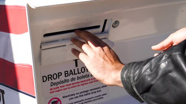 Young Man Drops Mail-in Ballot Letter in Slot at Official Voting Box with Drop Ballot Here Sign. Spanish Language Translation, Deposito de Boleta Aqui. For Immigrant Voters.