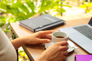 Mãos femininas segurando uma xicara de café branca em uma mesa de trabalho arrumada e organizada com um notebook e uma agenda com flores ao fundo