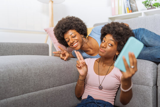 Twin sisters with afro hairstyle, posing while taking pictures with their cell phones, in the living room