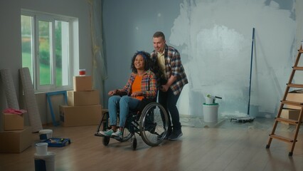Obraz na płótnie Canvas A white man and African American woman in a wheelchair. A young couple is planning a repair in the room. A room with a window, a ladder, cardboard boxes, wallpaper rolls and buckets of paint.