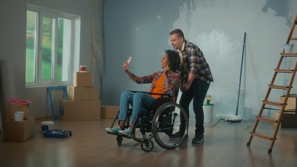 Obraz na płótnie Canvas A white man and an African American disabled woman in a wheelchair are talking on video call using a mobile phone. A young couple communicates and shares plans for repair the room.