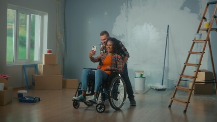 A white man and an African American disabled woman in a wheelchair are talking on video call using a mobile phone. A young couple communicates and shares plans for repair the room.