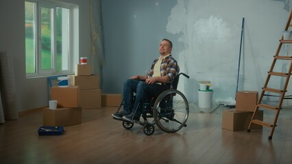 Fototapeta na wymiar A young man in a wheelchair. A disabled man thinks and plans repairs in the room. Handicapped person on the background of cardboard boxes, cans of paint and a ladder.