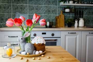 A bouquet of tulips, Easter bunnie, Easter cake and eggs on the table. In the background is a white Scandinavian-style kitchen.  The concept of home comfort and decor in the bright holiday of Easter.