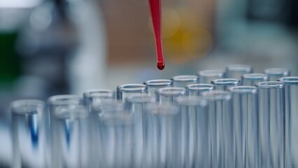 A drop of red liquid drips from a pipette into a transparent glass test tube. A dripping drop of blood and a row of test tubes with round necks close up. Sample analysis, research in laboratory.