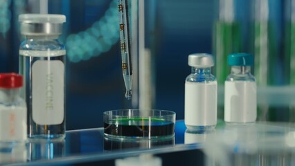 A drop of clear liquid drips from a glass pipette into a petri dish with a green substance. Nearby on the table are glass vials with a vaccine or medicine. Medical background, close up.