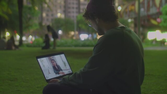 Man in casualwear and wireless earphones sitting on lawn in city park late in evening and speaking with female manager via online video call on laptop