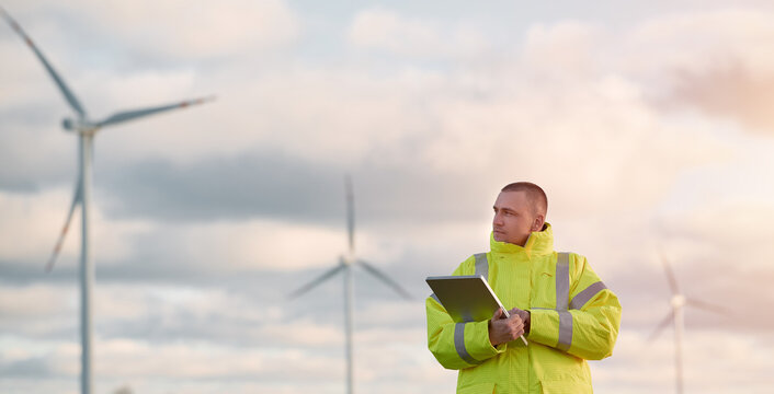 A male engineer man is standing in a wind turbine field with a beautiful sky background. Concept of sustainable future.