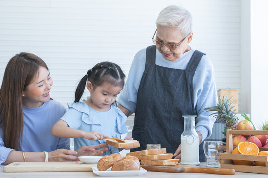 Happy Asian family preparing breakfast together. Grandmother and mother looking at child daughter girl spreading fruit jam on sliced bread and having fun in kitchen. Homemade food and little helper