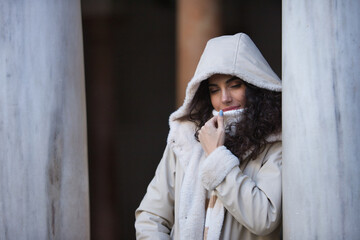 Young woman, beautiful, brunette with curly hair, sweater, with the coat hat on, leaning on marble columns, covering herself from the cold with the collar of the sweater. Concept autumn, winter, cold.