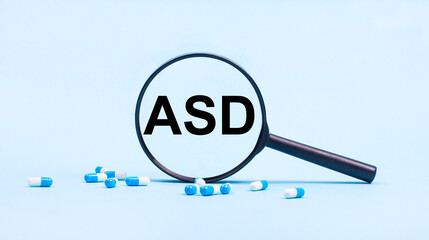 On a blue background, white and blue capsules with pills and a black magnifying glass with the text ASD Autism Spectrum Disorder. Medical concept.