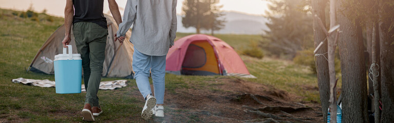 Rear on couple walking at camping place hand in hand and carrying portative fridge.