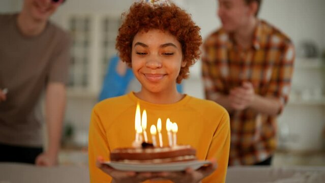 Portrait smiling african american birthday girl with cake in hands looks at burning candles makes a wish. Happy party with friends clapping hands and blowing horns on background. Celebration concept.