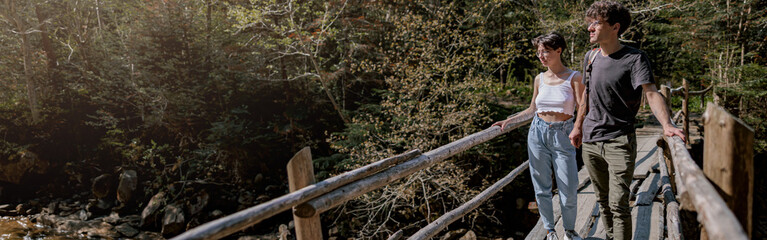 Obraz premium Couple of hikers crossing river by wooden bridge in mountains. Fast water. Trekking.