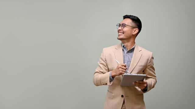 Smiling Asian Businessman Looking Aside At The Empty Space, Holding A Tablet