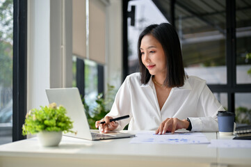 Focused Asian businesswoman using laptop, analyzing financial data, working at her desk.