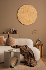 Warm and cozy living room interior with round wall panel, stylish sofa, brown plaid, beige coffee table, vase with dried flowers, books, modern sculpture and personal accessories. Home decor. Template