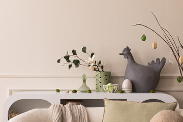 Creative composition of easter living room interior with copy space, hen sculpture, easter eggs, easter bunny, white sofa, beige pillows and personal accessories. Home decor. Template.