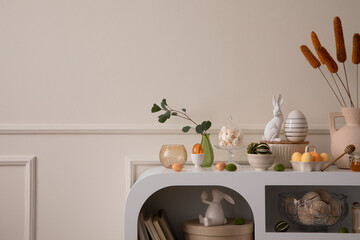Minimalist composition of easter living room interior with copy space, easter bunny sculpture, easter eggs, vase with dried flowers, wall with stucco and personal accessories. Home decor. Template.