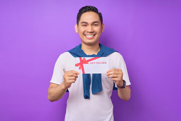 Smiling young Asian man in casual clothes holding a gift certificate, looking at the camera...