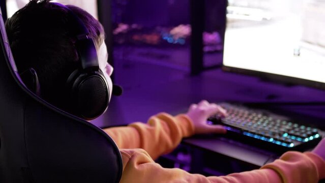 Boy Gamer Playing Video Game, Sitting at a Computer in a Virtual Room at Night. Cybersport club virtual reality. E-sport competition. Headset, neon lighting, keyboard, mouse. VR. Screen. Gambling.