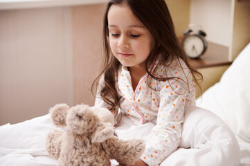Adorable elementary age Caucasian child girl in stylish pajamas, sitting on bed after waking up in the morning, playing with a plush toy sheep, in her cozy bedroom. Children. Happy childhood concept