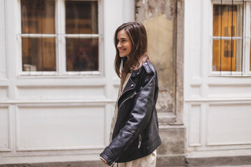 Obraz premium Trendy girl with brunette hairstyle posing outside. Woman in leather jacket and shirt walking on the street outdoors. Tourist happy woman posing in the city. Optimistic lady turn around.