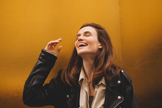 Simpering young brunette woman with a coy smile as she flirts over a gold studio background with copy space. Woman wear black leather jacket and beige shirt is laughing.