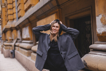 Trendy girl with blonde hairstyle posing outside. Woman in grey jacket and hoodie walking on the street outdoors. Tourist happy woman posing in the city. Optimistic lady put on hood walks outside.