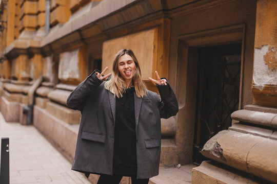 Cool blonde woman in black hoodie and grey jacket shows peace sign, show tongue, close eyes and posing outside. Happy woman walk and posing outdoor.