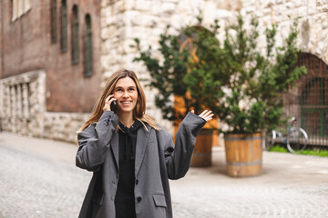 Young caucasian attractive smiling blonde woman in a grey jacket talking on a mobile phone flirting and twisting a lock of hair on her finger walking outside. Girl flirting.