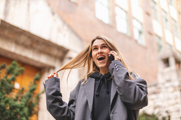 Surprised young caucasian attractive smiling blonde woman in a grey jacket talking on a mobile phone hear good news, open mouth and twisting a lock of hair on her finger walking outside. Girl flirting