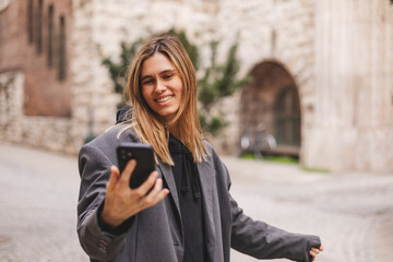 Happy young woman photographing herself using her mobile phone. Caucasian female talking selfie with her smart phone walking outdoor on the street. Girl posing in grey suit, t-shirt.