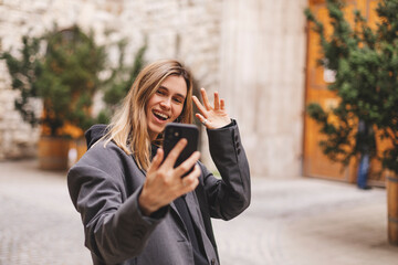 Woman waiving saying hello happy and smiling, friendly welcome gesture. Portrait of young beautiful attractive smiling cheerful girl taking selfie saying hello outside outdoors.