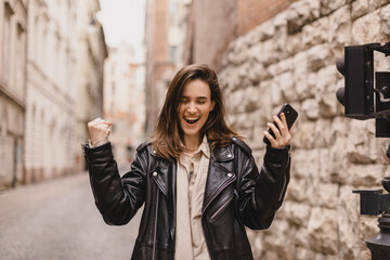 Delighted brunette girl making winner gesture and using mobile phone walking on the street. Portrait of surprised hipster girl surfing internet on mobile phone outdoors.